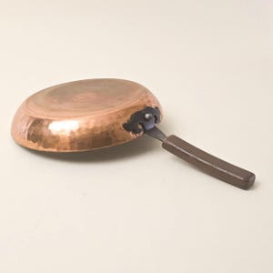 Peut inclure: Une poêle de couleur cuivrée avec une texture martelée. La poêle a un manche marron foncé et une fixation métallique. La poêle est ronde et semble être vintage.