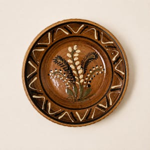 May include: A round ceramic plate with a glossy brown glaze. The center features a hand-painted floral motif in cream, dark green, and black. The rim is decorated with a repeating pattern of cream and black V-shaped lines and small dots.