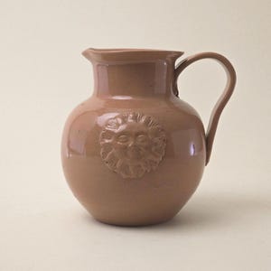 Könnte beinhalten: Ein hellbrauner Keramikkrug mit rundem Körper und gebogenem Henkel. Der Krug hat ein erhabenes Sonnengesicht-Design auf der Vorderseite. Der Ausguss ist leicht ausgestellt. Dieser dekorative Krug eignet sich zum Servieren von Getränken.