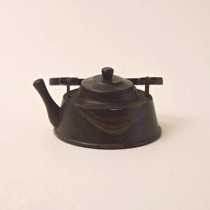 May include: A dark brown wooden teapot featuring a smooth, rounded body with a visible wood grain pattern, a curved spout, a flat lid with a small central knob, and a horizontal handle attached to the top with two vertical supports.