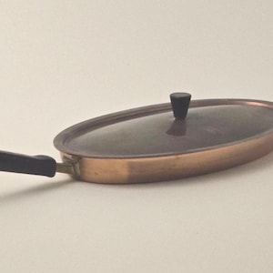 May include: Oval copper-colored pan with a black handle and a clear lid with a black knob. The pan is designed for cooking and has a classic, vintage aesthetic. The handle is angled for easy grip.