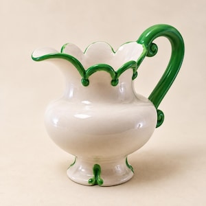 May include: A cream-coloured ceramic pitcher featuring a glossy finish, a scalloped rim outlined in vibrant green, and a matching green curved handle. The base of the pitcher is also decorated with green accents, creating a cohesive design.