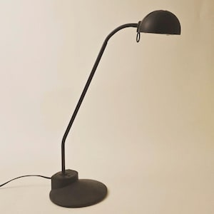 May include: A matte black desk lamp featuring a circular base, a thin adjustable neck, and a dome-shaped shade. The lamp includes a small pull-ring attached to the underside of the shade and a power cord extending from the back of the base.