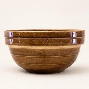 May include: A brown ceramic mixing bowl with a glossy finish and a wide, reinforced rim. The exterior features three horizontal ridges encircling the body, with a slightly tapered base and a smooth, rounded interior surface for food preparation.