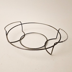 May include: A dark metal serving basket with a circular base and oval top. The handles are wrapped with a black and white twisted design. The basket is designed to hold a round dish.