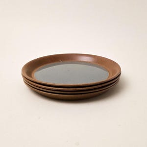 May include: A stack of four small, round ceramic plates. The plates have a brown exterior and a light blue-grey interior. The plates are stacked, with the top plate slightly angled.