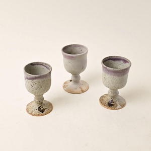 Puede incluir: Tres copas de cerámica hechas a mano. Cada copa tiene un cuerpo moteado blanco roto con un borde morado. Las bases son redondas y moteadas, con un acento marrón más oscuro. Las copas están dispuestas sobre un fondo claro.
