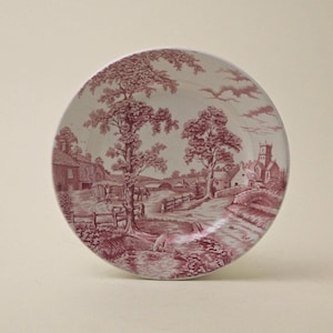 May include: A pink and white ceramic plate with a countryside scene. The plate features a farmhouse, trees, a bridge, and a church in the background.