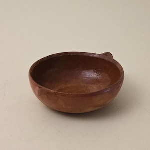 Puede incluir: Un pequeño cuenco de terracota marrón con una sola asa. El cuenco es ligeramente redondeado y tiene una superficie lisa y ligeramente texturizada.