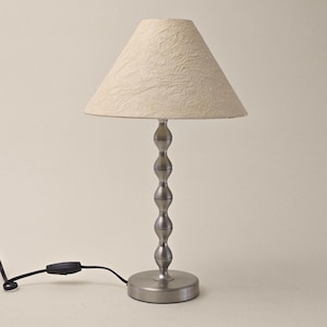 May include: A small table lamp with a cream-coloured, textured lampshade. The lamp has a silver-coloured, beaded stem and a round base. The lamp is connected to a black power cord with an on/off switch.