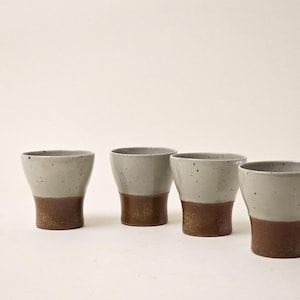 Puede incluir: Cuatro tazas de cerámica con un diseño de dos tonos. La parte superior es gris moteado y la parte inferior es marrón oscuro. Las tazas tienen una forma ligeramente acampanada y un acabado liso. Están dispuestas en fila.