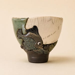May include: A ceramic cup featuring a textured, dark brown base and a two-toned upper section. One side is a smooth, cream-colored crackle glaze, while the other is a textured green. A sculpted, dark gray fish with a white eye is attached to the side.