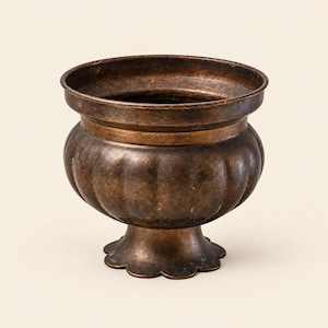 Peut inclure: Un pot en métal de couleur bronze foncé avec un bord large et évasé et un corps arrondi et nervuré. Le récipient repose sur une base en piédestal avec un bord festonné. La surface présente une finition mate patinée avec des nuances.