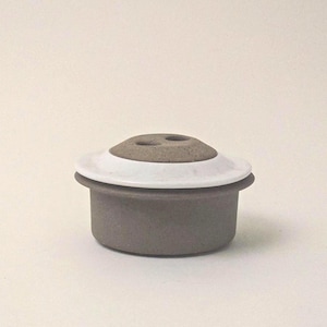 May include: A small, gray ceramic container with a white rim and a rounded, light brown top. The top has two small holes. The container is likely for holding small items or as a decorative piece.