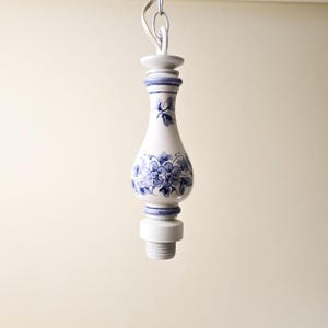 May include: A white ceramic pendant light fixture with blue floral designs. The fixture has a bulbous shape with a narrow neck and a threaded base. The light is suspended by a white chain.
