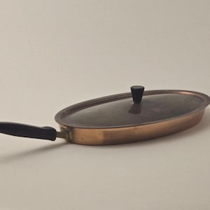 Puede incluir: Una sartén ovalada de cobre con un mango negro y un pomo negro en la tapa.