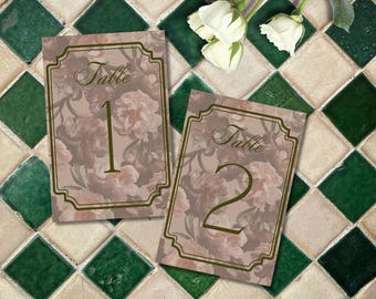 May include: Two floral table number cards with gold frames. The cards are on a green and white tiled surface. The first card says "Table 1" and the second card says "Table 2".
