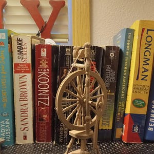 May include: A wooden spinning wheel with a white spindle and a brown wooden base. The spinning wheel is sitting on a shelf with a row of books behind it.