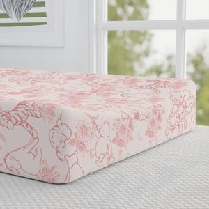 Puede incluir: Una sábana bajera ajustable para cuna con un estampado floral y de personajes en rosa y blanco. El diseño presenta ilustraciones de un oso, un tigre y otros personajes, junto con flores y árboles. La sábana es de tela suave.