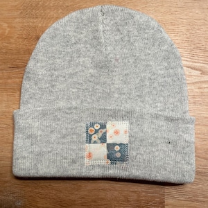 May include: A grey beanie hat with a turned-up brim. The hat has a square patch on the front, divided into four sections with floral and geometric patterns in blue, white, and peach.
