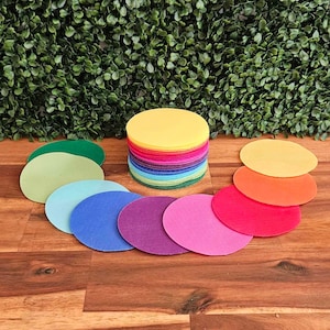 Pode incluir: Uma pilha de círculos de feltro coloridos em vários tons de verde, azul, roxo, rosa, laranja e amarelo. Os círculos estão dispostos em semicírculo sobre uma superfície de madeira.