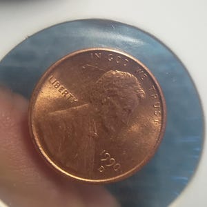 Może przedstawiać: Miedziana moneta 1-centowa Stanów Zjednoczonych z 1990 roku, z profilem Abrahama Lincolna. Moneta jest zamknięta w ochronnym uchwycie. Widoczne są słowa "LIBERTY" i "IN GOD WE TRUST".