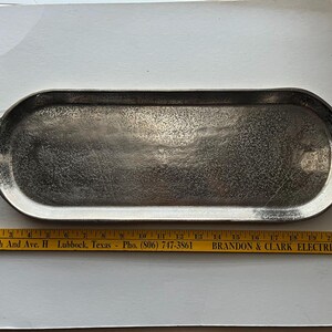 Puede incluir: Una bandeja de servir de metal plateado con extremos redondeados y asas de bucle. La bandeja tiene una superficie texturizada y mide aproximadamente 51 cm de largo. Una regla amarilla con texto est&aacute; debajo.