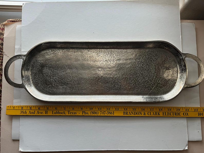 Puede incluir: Una bandeja de servir de metal plateado con extremos redondeados y asas de bucle. La bandeja tiene una superficie texturizada y mide aproximadamente 51 cm de largo. Una regla amarilla con texto est&aacute; debajo.
