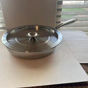 May include: A stainless steel saucepan with a lid. The pan has a long handle and a small, rounded knob on the lid. The pan is sitting on a white surface.