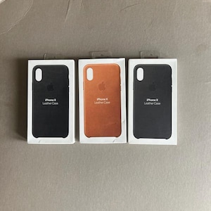 以下が含まれることがあります： iPhone X用レザーケースが入った3つの白い長方形の小売用ボックス。2つのボックスには黒いケース、1つのボックスにはタンカラーのレザーケースが入っています。各ボックスにはAppleのロゴと「iPhone X Leather Case」というテキスト、カメラ用の切り抜きが表示されています。