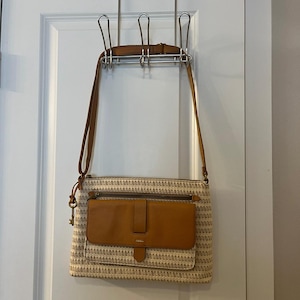 May include: A beige and brown crossbody bag with a woven pattern and a brown leather flap pocket. The bag has a long brown strap and a key-shaped charm. It is hanging on a silver metal door hanger.