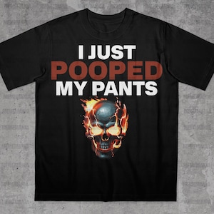 Peut inclure: T-shirt noir avec l'inscription "I JUST POOPED MY PANTS" en blanc et rouge. Un cr&acirc;ne en flammes avec des yeux jaunes est plac&eacute; sous le texte. Le cr&acirc;ne est entour&eacute; de flammes orange.