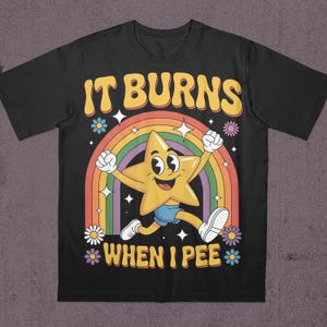 May include: Black t-shirt with a retro design featuring a smiling yellow star character, a rainbow, and the text "IT BURNS WHEN I PEE". The design includes flowers and stars.