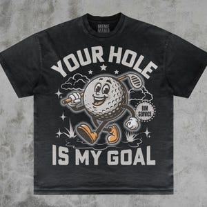 Puede incluir: Camiseta gris oscuro con un personaje de bola de golf de dibujos animados sosteniendo un palo de golf. La camiseta presenta el texto "YOUR HOLE IS MY GOAL" en blanco, con el logo "Meme Mania" en la parte superior.