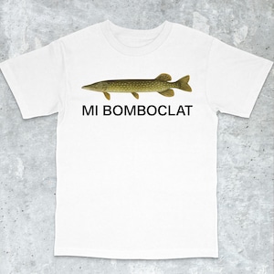 Könnte beinhalten: Weißes T-Shirt mit einer detaillierten Illustration eines Fisches in Grün- und Brauntönen. Unter dem Fisch ist der Text "MI BOMBO CLAT" in schwarzen, fetten Buchstaben aufgedruckt. Das T-Shirt hat einen klassischen Rundhalsausschnitt.