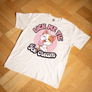 Op de afbeelding: Een wit T-shirt met een roze cirkel en een cartoonkat die een ijsje likt. De tekst "Lick Me Til Ice Cream" staat rond de cirkel.