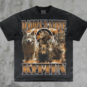 May include: Dark grey t-shirt with a graphic of wolves and a fiery background. The text "DADDY'S LIL' KITTEN" is printed in gold. The shirt has a vintage, distressed appearance. The brand name "MEME MANIA" is printed on the collar.