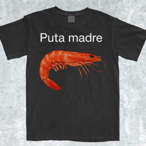 May include: Black t-shirt featuring a large, vibrant red shrimp illustration. Above the shrimp, the text "Puta madre" is printed in white. The shirt is made of a soft material.