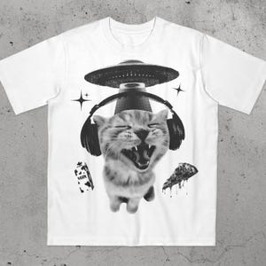 Pode incluir: Camiseta branca com um gráfico em preto e branco de um gato usando fones de ouvido e um chapéu de OVNI. O gato está sorrindo com a boca aberta. Uma fatia de pizza e uma caixa de leite também estão no design.