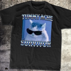 May include: Black t-shirt featuring a white cat wearing sunglasses and the text "Tummy Ache Survivor" in a blue box.