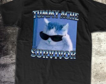 May include: Black t-shirt featuring a white cat wearing sunglasses and the text "Tummy Ache Survivor" in a blue box.