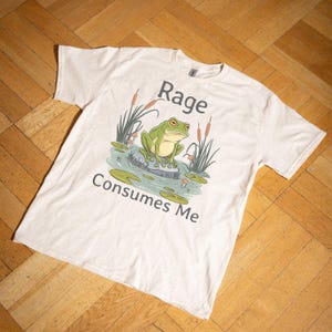 Puede incluir: Una camiseta beige con una rana verde sentada en una roca en un estanque. El texto "Rage Consumes Me" está impreso en la camiseta.