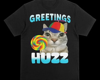 May include: A black t-shirt featuring a graphic of a cat wearing a multicolored propeller hat. The cat is holding a rainbow-colored lollipop. The text GREETINGS is printed in light blue above the cat, and the word HUZZ is printed in white below.