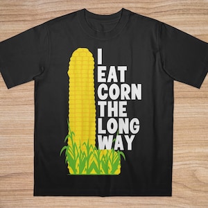 Op de afbeelding: Zwart T-shirt met een gele maïsafbeelding en groen gras. Witte tekst luidt "I EAT CORN THE LONG WAY". Het ontwerp toont een verticale maïskolf illustratie, die een humoristische uitspraak doet.