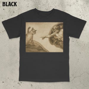Puede incluir: Camiseta negra con un gráfico humorístico. El diseño presenta un gato y una representación de la Creación de Adán de la Capilla Sixtina. El gato mira la mano de Dios. La palabra "BLACK" está impresa en la parte superior.