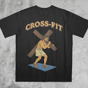Può includere: T-shirt nero con la scritta "CROSS-FIT" in un font color beige a blocchi. La maglietta presenta un'illustrazione a cartoni animati di un uomo che porta una croce, con una corona di spine e pantaloncini.