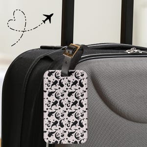 Może przedstawiać: Prostokątna etykieta bagażowa z czarnym skórzanym paskiem i złotą klamrą jest przymocowana do czarnej walizki. Etykieta przedstawia wzór czarnych syren, nut muzycznych i księżyców na białym tle. Przerywana linia z sercem i samolotem znajduje się w tle.