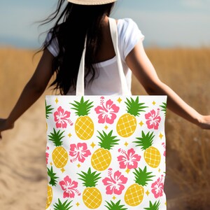 Può includere: Borsa tote bianca con un motivo tropicale di ananas gialli, fiori di ibisco rosa, foglie verdi e accenti di diamanti gialli. La borsa ha una tracolla bianca.