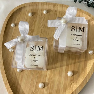 May include: Two clear plastic boxes with white satin bows and pearl accents. Each box contains two white sweets and a printed card with the initials "S/M", the names "Sedanur & Mert", and the date "11.01.2025".