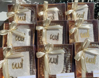 May include: A collection of small, clear gift boxes with white fabric and gold lettering inside. Each box contains a white pearl necklace and a small white fabric square with gold Arabic calligraphy. The boxes are tied with white ribbon and have a gold border. The text on the boxes reads "Mashallah Iznah & Noah".